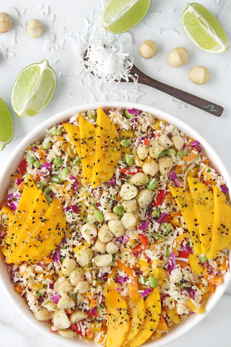 Fresh and colorful tropical hearts of palm rice salad with coconut lime flavors, colorful vegetables, and a macadamia nut garnish, shown with an overhead perspective on a white marble background with lime wedges.