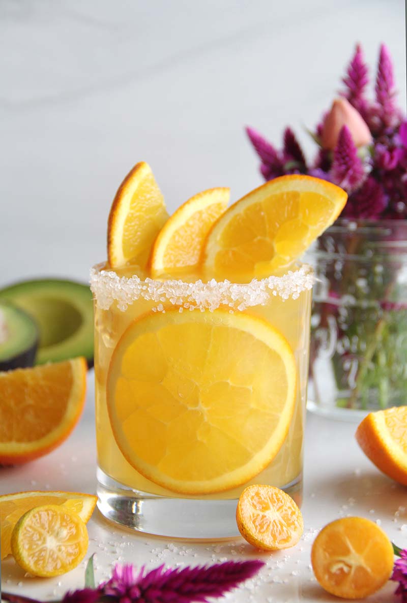 Cozy spiced orange margarita made with fresh orange juice, tequila, and spiced rum, garnished with a salted rim and orange slices, shown in a bright kitchen with purple flowers.