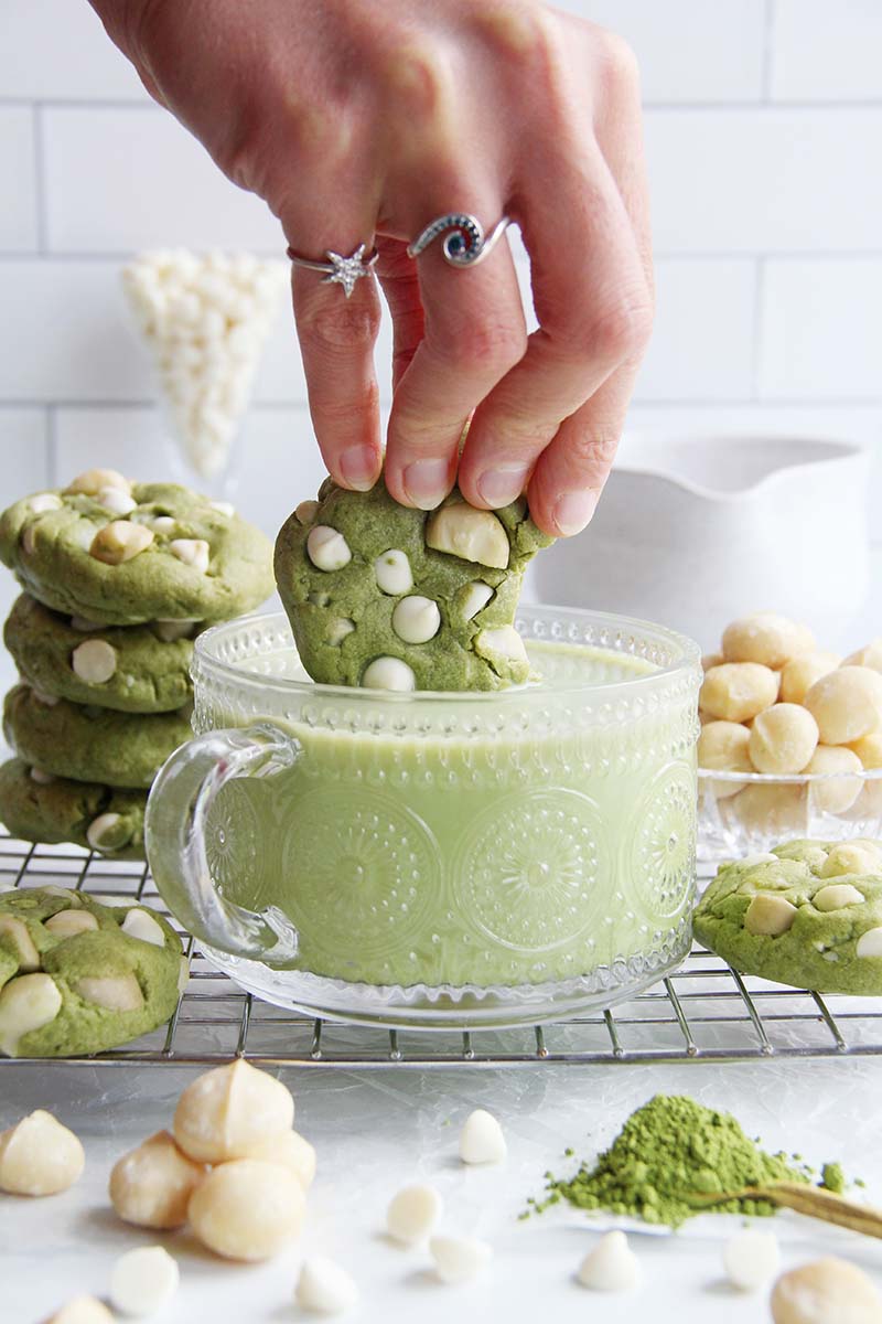 Vegan matcha cookie with white chocolate and macadamia nuts held in a woman's hand and being dipped into a creamy matcha latte.