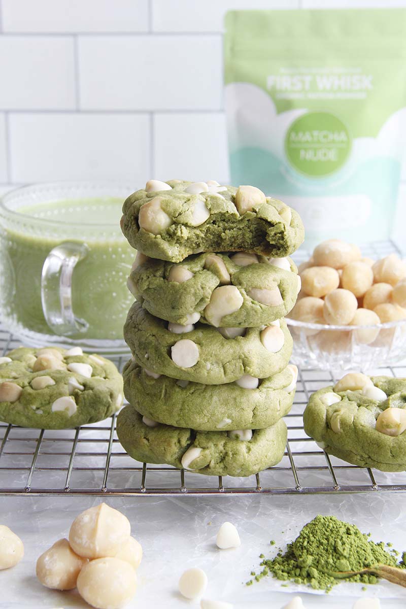 Stack of vegan matcha cookies with white chocolate chips and macadamia nuts, shown on a cooling rack with a bag of matcha powder in the background.