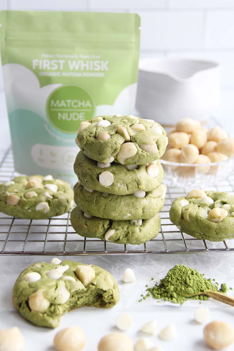 Vegan white chocolate matcha cookies stacked on a cooling rack with a bag of matcha powder in the background and macadamia nuts in the foreground.