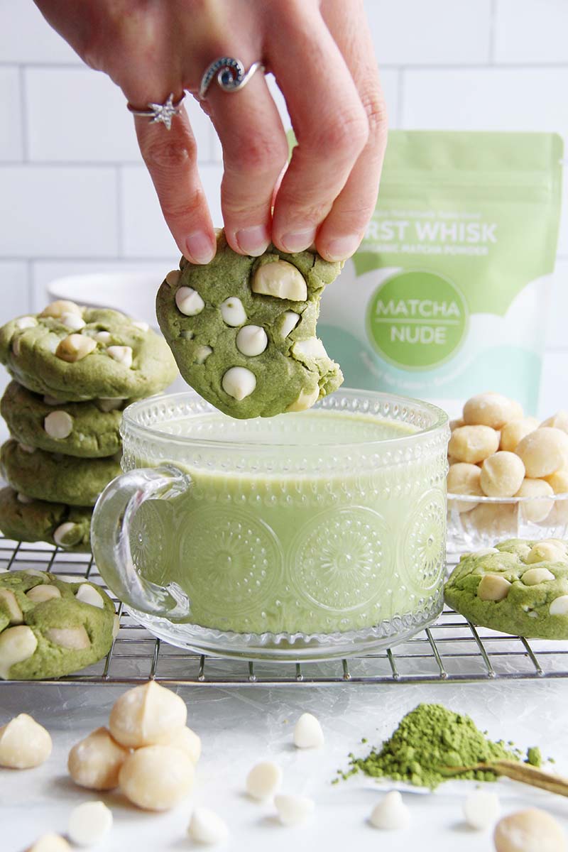 Hand dipping a gluten-free matcha cookie into a latte, with stacked cookies and a bag of matcha powder in the background.
