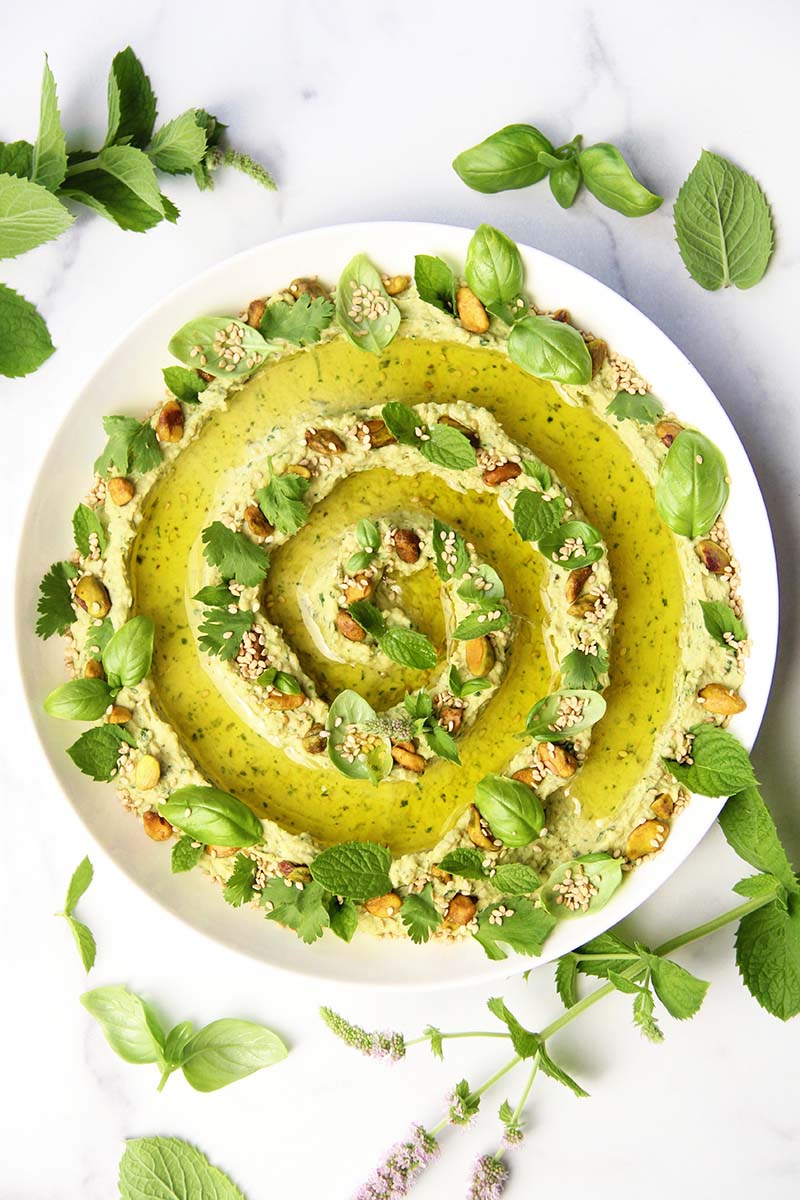 Green goddess hummus garnished with fresh herbs, pistachios, sesame seeds, and olive oil, shown on a white surface alongside herb sprigs.