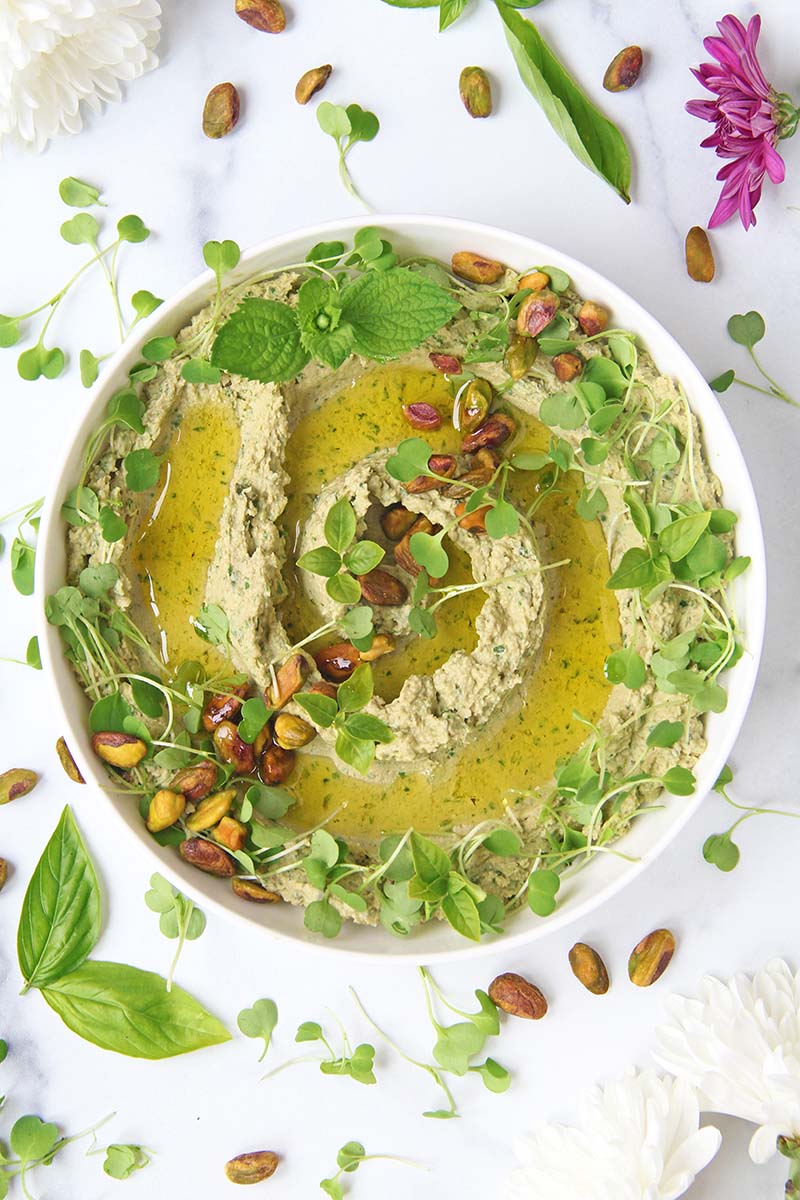 Bowl of green goddess hummus topped with pistachios and microgreens, shown in a white kitchen alongside spring flowers.
