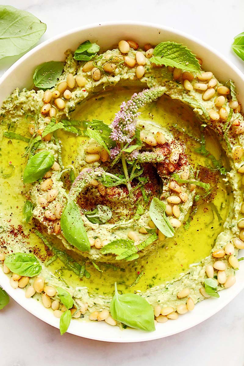 Close-up of a bowl of green goddess hummus with topped with pine nuts, sumac, and mint flowers.