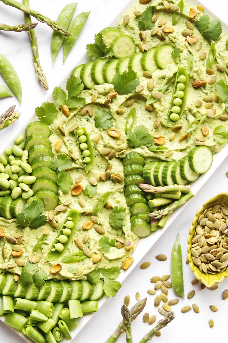 Green goddess hummus platter topped with cucumbers, peas, asparagus, and green onions, ready for a spring party.