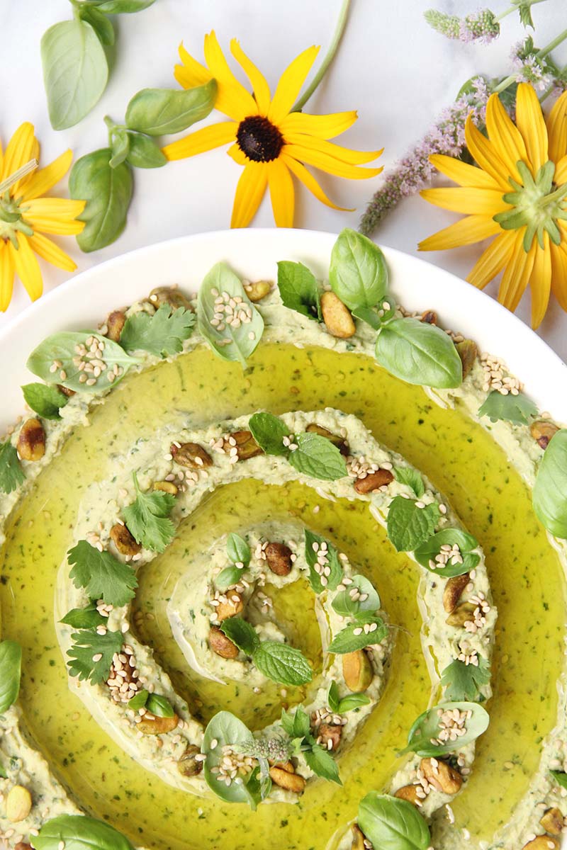Bowl of fresh herb green goddess hummus, garnished with sesame seeds, pistachios, and herbs, shown alongside yellowflowers and herb sprigs.