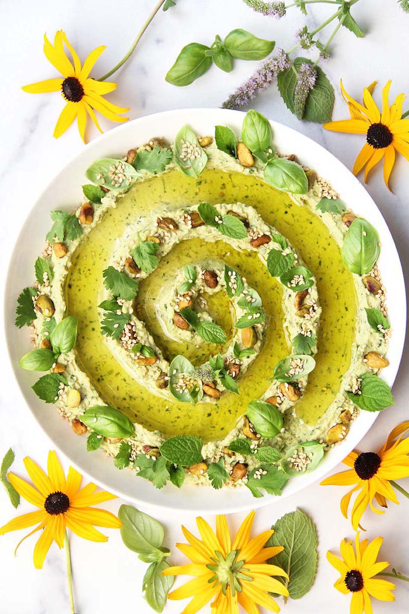 Vibrant green goddess hummus with fresh herbs, sesame seeds, and pistachios, shown in a serving bowl on a white marble countertop.