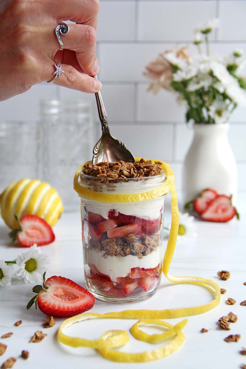 Hand holding a spoon dipping into a strawberry yogurt parfait; the parfait is shown in a glass jar and layers of granola, yogurt, and strawberries are visible.