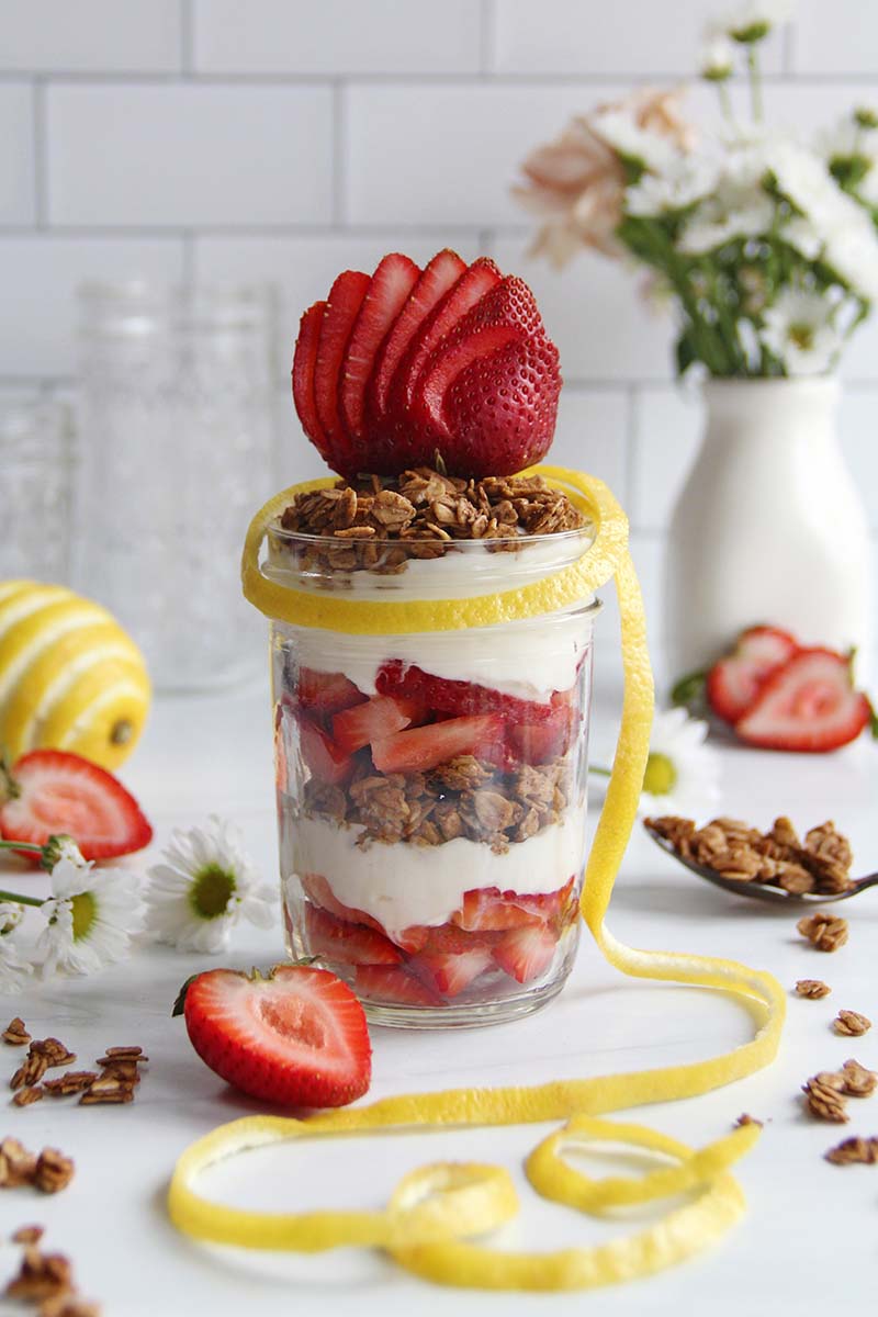 Healthy strawberry lemon yogurt parfait layered with granola and topped with strawberry slices, styled with fresh berries and white spring flowers.