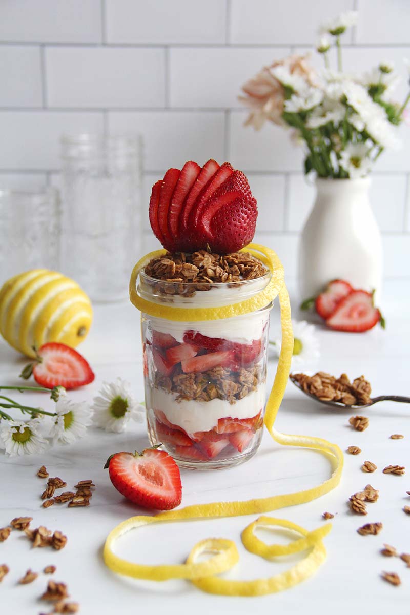 Layered strawberry yogurt and granola parfait in a glass jar, surrounded by fresh strawberries and white flowers for a spring breakfast setting.
