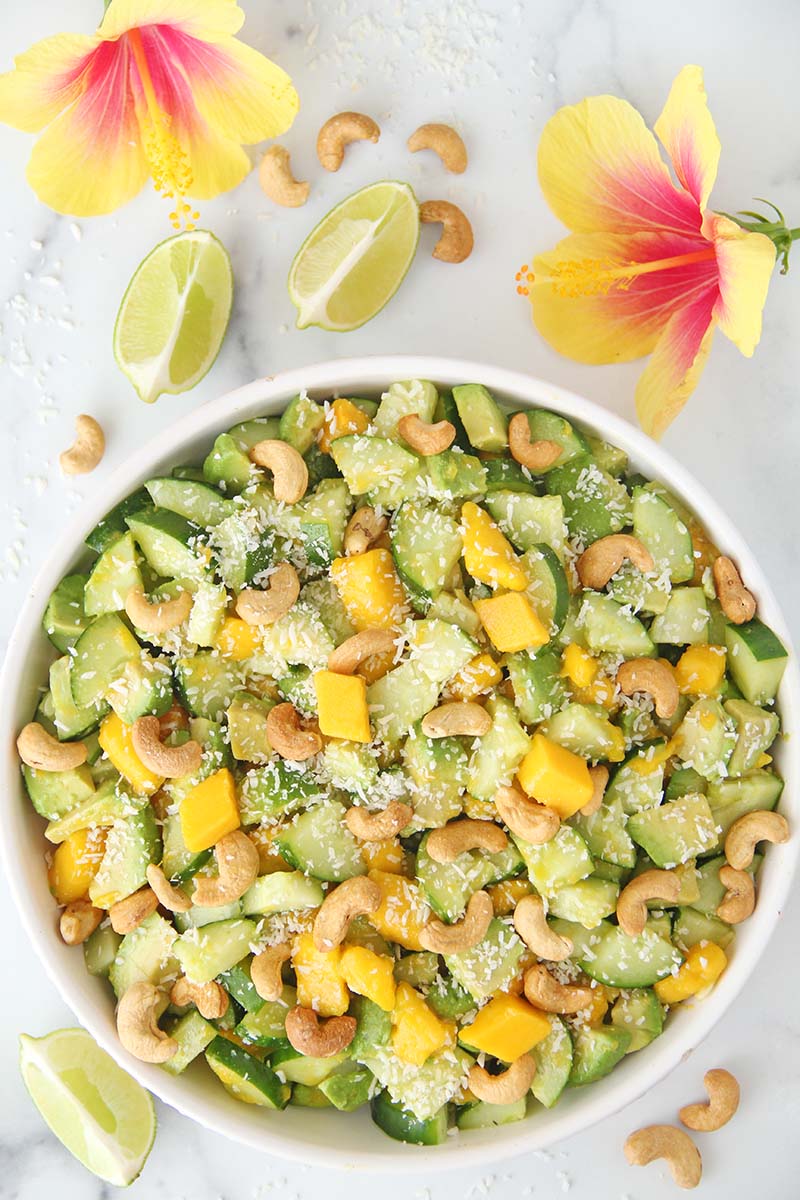 Overhead view of a bowl of Mango Cucumber Avocado Salad in a white kitchen with tropical flowers.