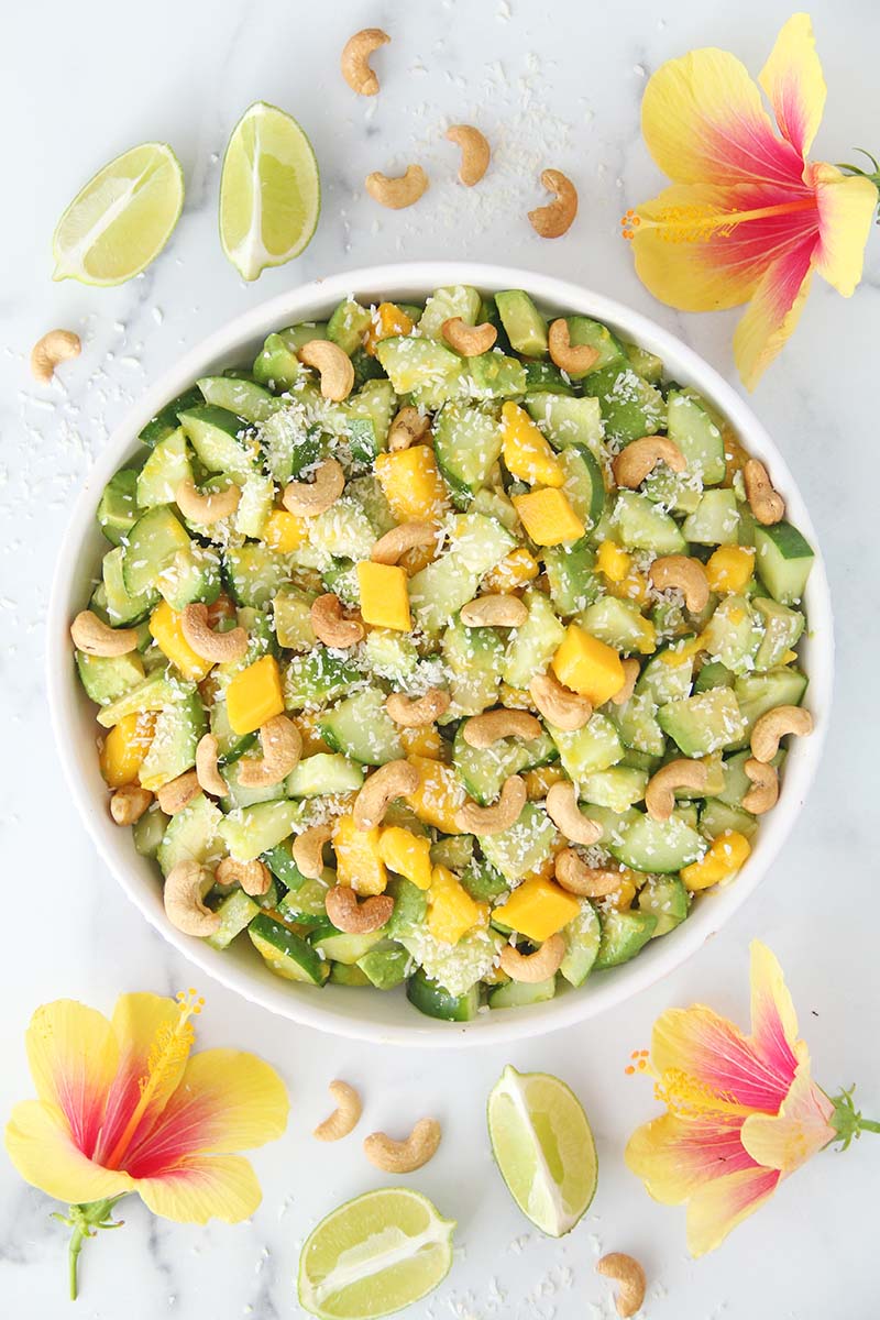 Bowl of Mango Cucumber Avocado Salad, shown alongside tropical flowers, lime slices, and extra cashews.