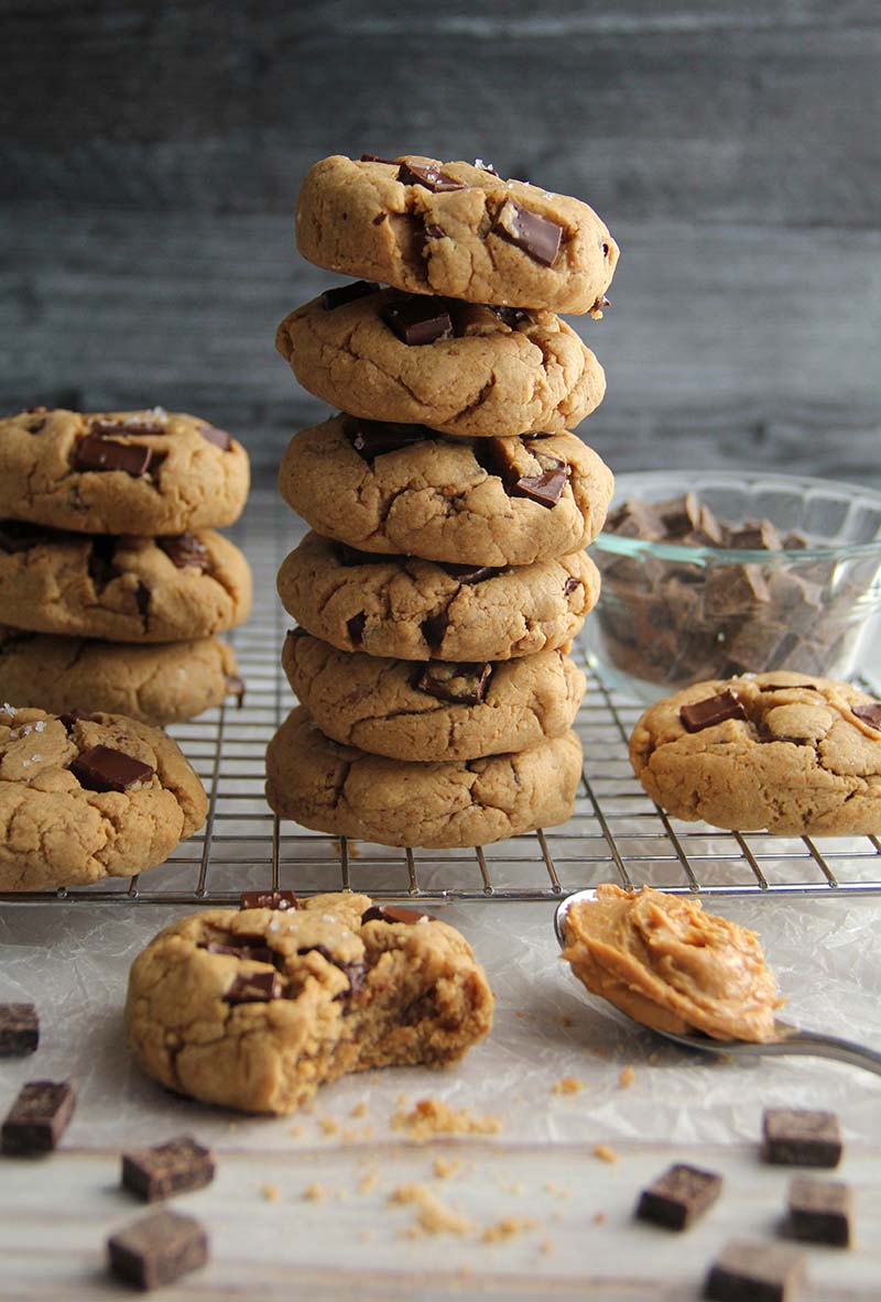 Stack of vegan gluten-free peanut butter chocolate chunk cookies on a cooling rack with scattered chocolate chunks and a spoonful of peanut butter in the foreground.