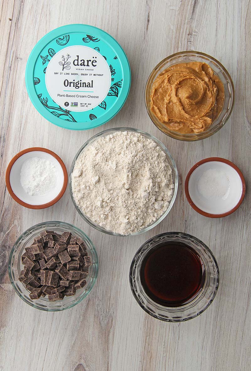 Overhead view of ingredients for vegan gluten-free peanut butter chocolate chunk cookies, including oat flour, peanut butter, maple syrup, and vegan cream cheese.