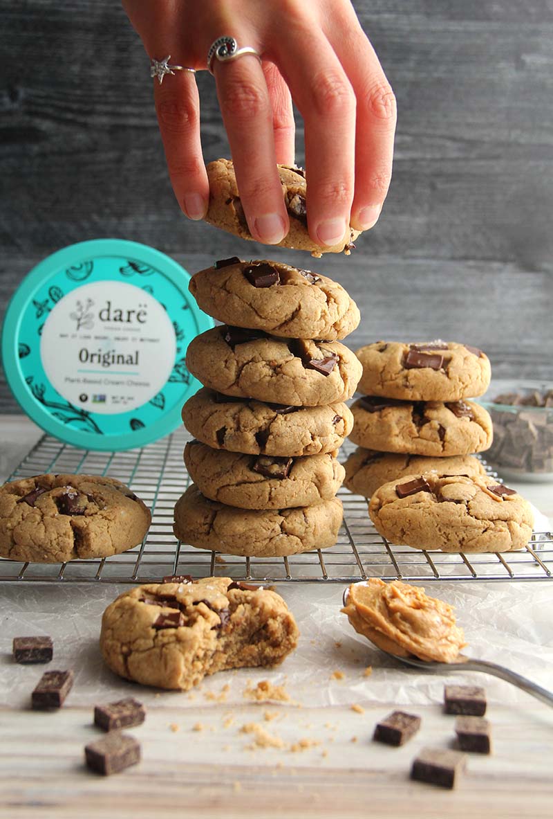 Hand reaching for a vegan gluten-free peanut butter chocolate chunk cookie, shown with chocolate chunks, peanut butter, and vegan cream cheese in the background.