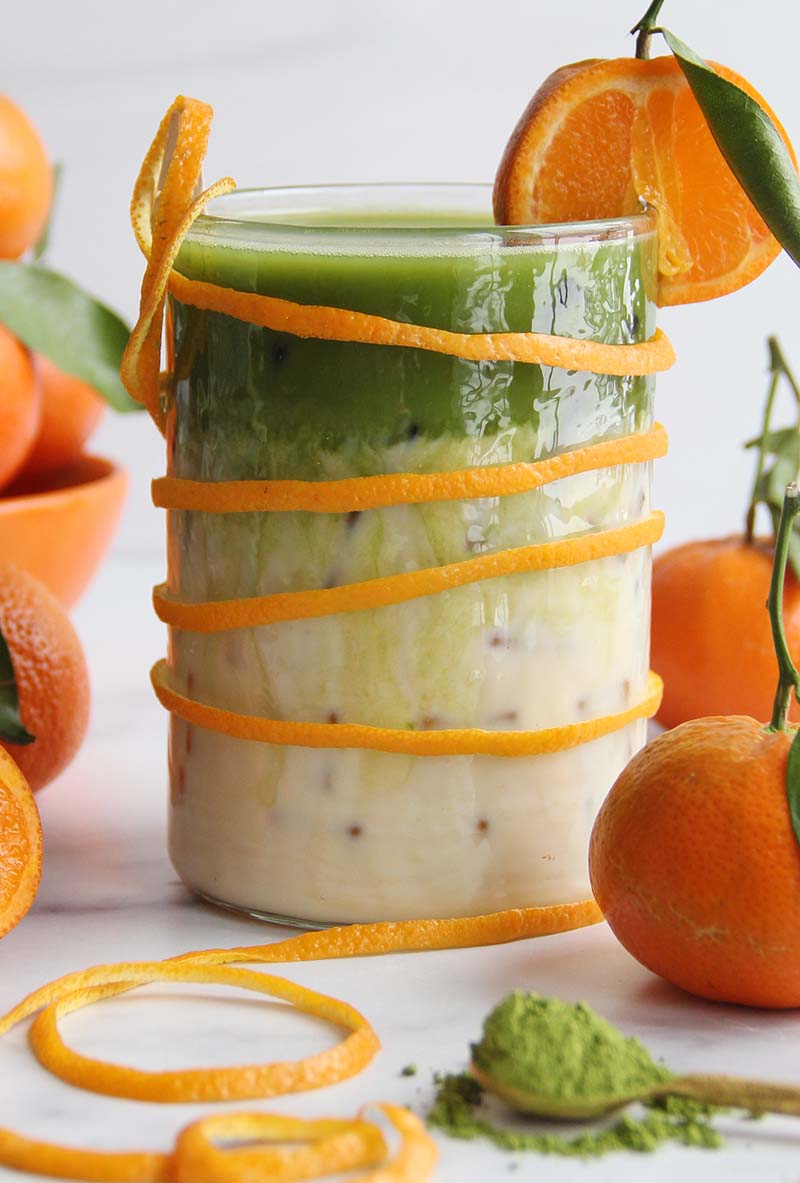 Close-up of an iced orange blossom matcha latte in a glass, garnished with an orange slice and orange peel twist, with fresh orange slices and matcha powder in the foreground.