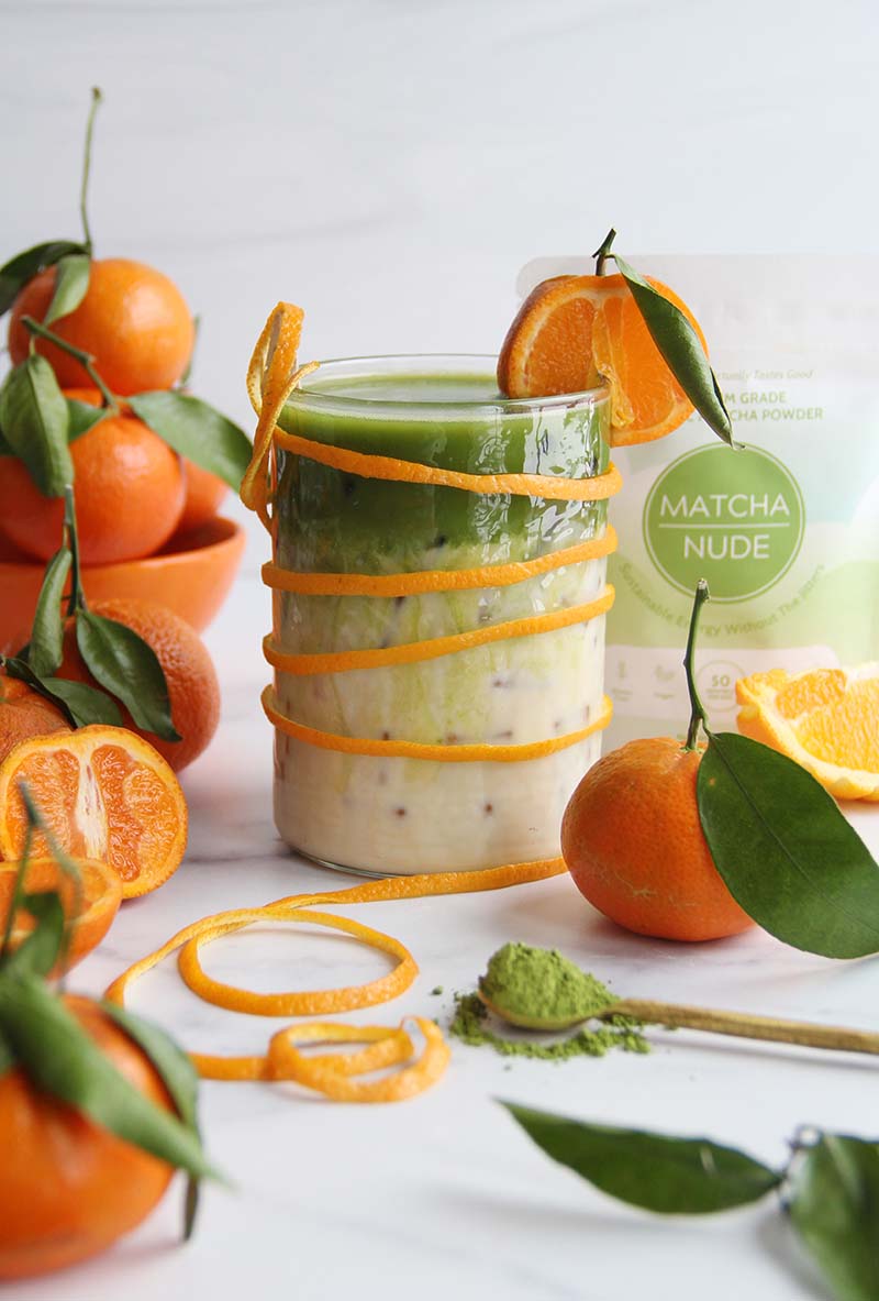 Iced orange blossom matcha latte in a white kitchen, shown with a bag of matcha in the background and orange slices and matcha powder in the foreground.