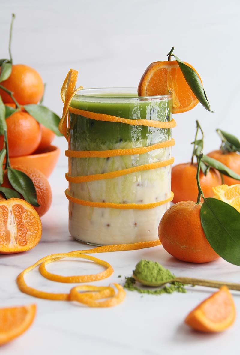 Citrusy orange blossom matcha latte over ice in a glass, garnished with an orange slice and peel, shown with fresh oranges and matcha powder.