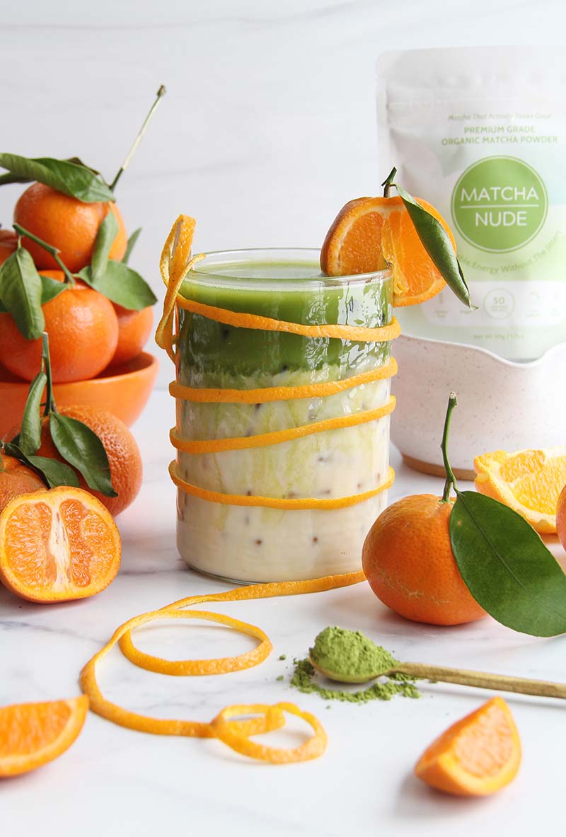 Glass of iced orange blossom matcha latte with an orange slice garnish, shown with a matcha bag in the background, and sliced oranges and matcha powder in the foreground.