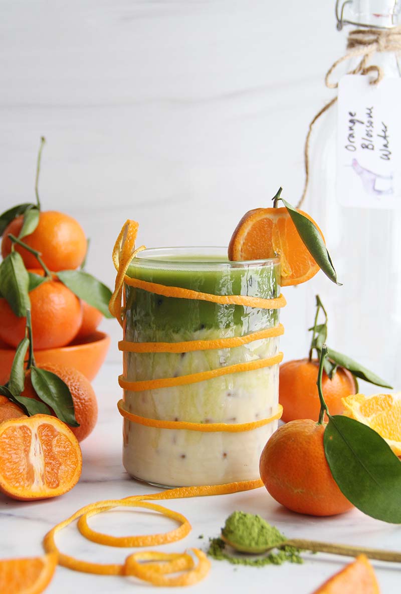 Iced orange blossom matcha latte with orange slice garnish, shown with a bottle of orange blossom water in the background, and fresh oranges and matcha powder in the foreground.
