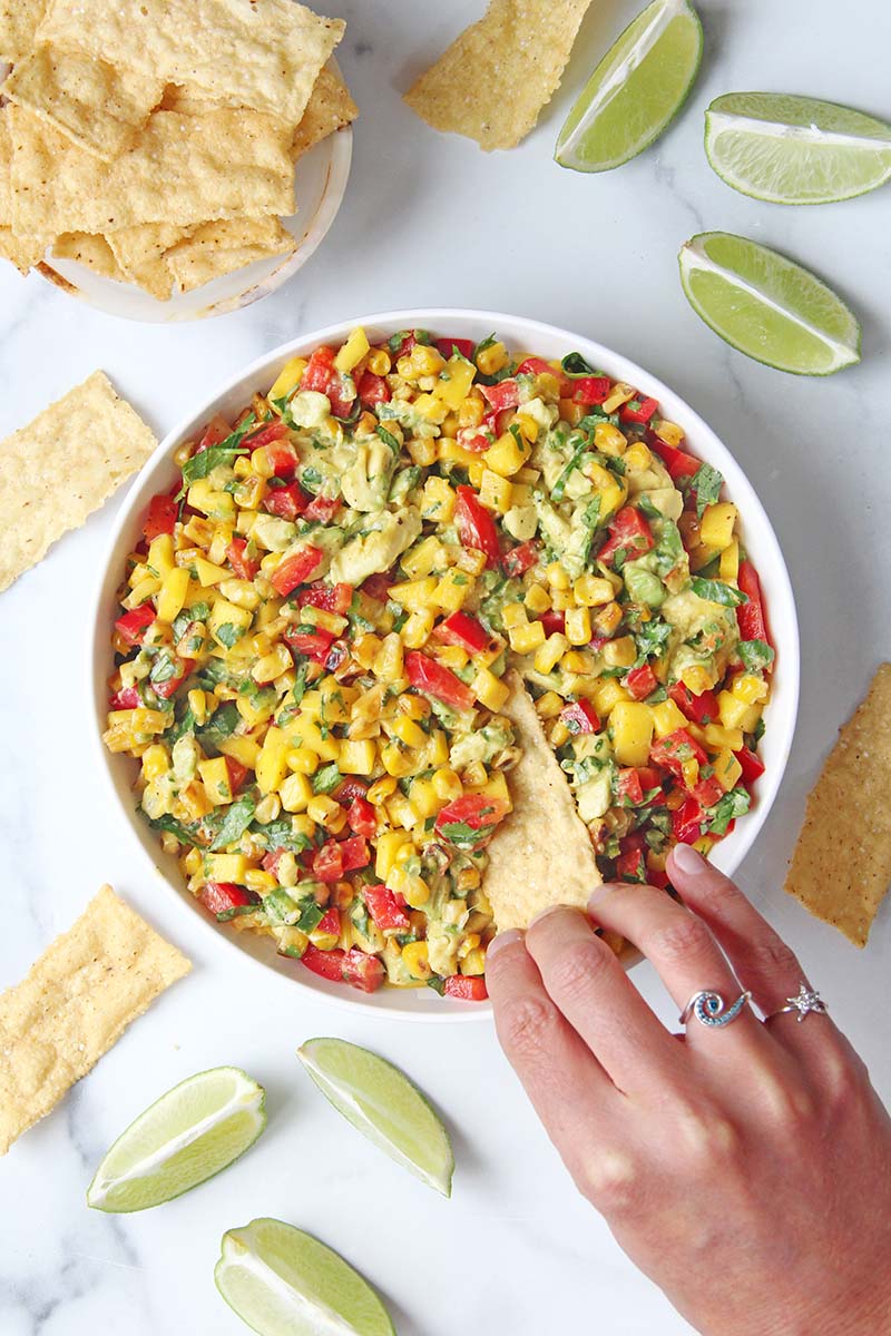 Woman's hand dipping a tortilla chip into sweet and spicy corn and mango salsa, shown in a white kitchen next to a bowl of chips.