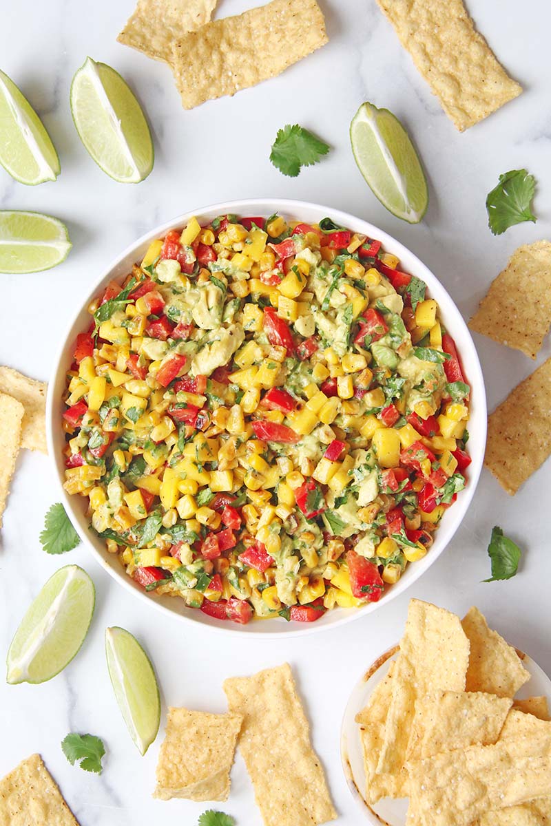 Tropical corn and mango salsa in a serving bowl, shown with tortilla chips and lime wedges in a white kitchen.