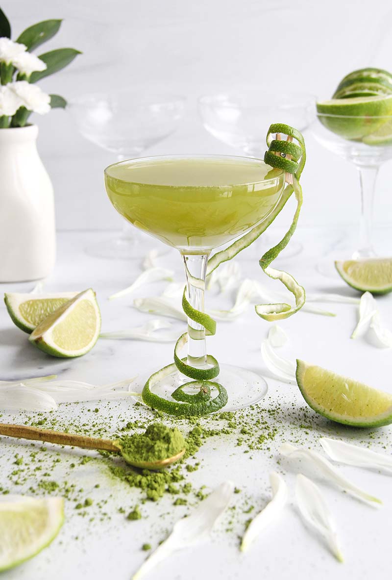 Green matcha daiquiri cocktail in a coupe glass with curly lime peel, shown with white flowers and extra lime wedges in a white kitchen.