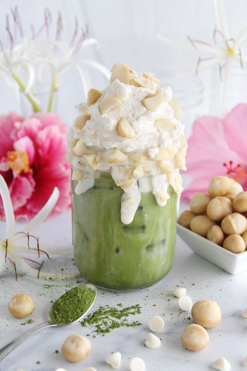 White chocolate macadamia matcha latte, with a spoonful of matcha in the foreground and Hawaiian flowers in the background.