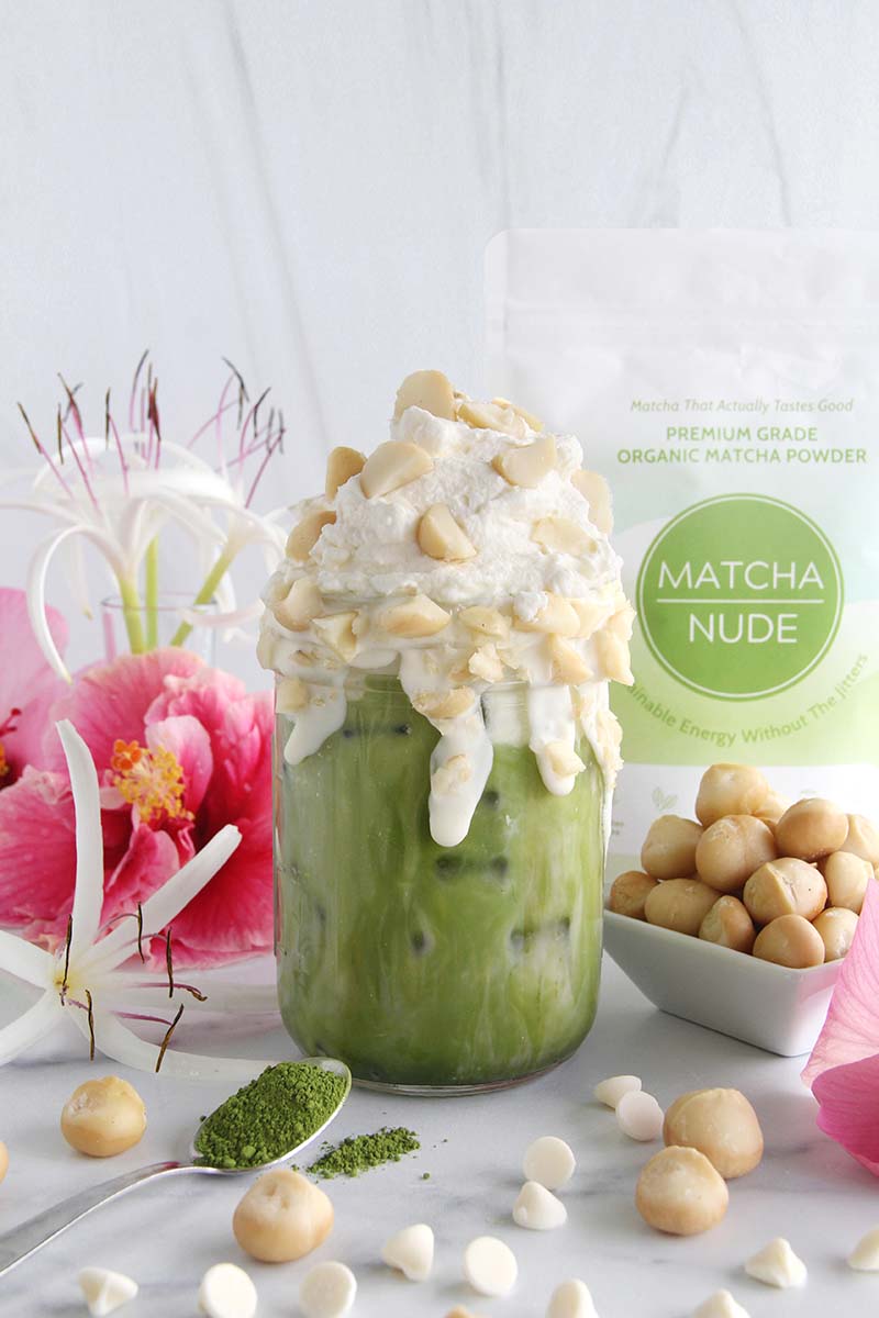 White chocolate macadamia matcha latte topped with whipped cream and macadamia nuts, shown with a bag of matcha in the background.