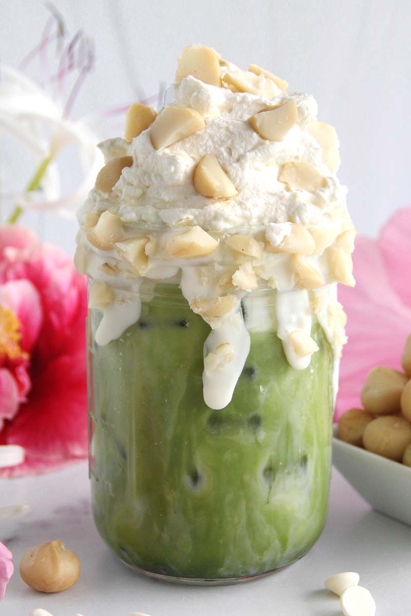 Close-up of a white chocolate macadamia matcha latte topped with whipped cream and crushed macadamias, shown with Hawaiian flowers.