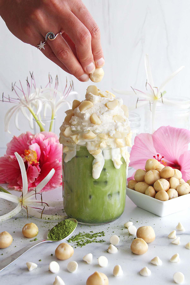 Woman's hand adding macadamia nuts on top of a white chocolate macadamia matcha latte, shown in a white kitchen.