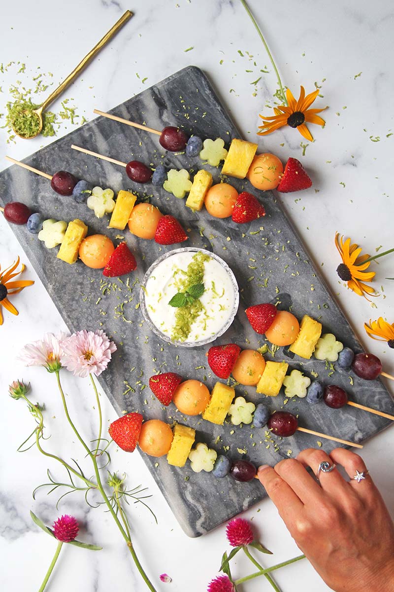 Overhead view of rainbow fruit skewers on a stone serving platter, with a bowl of mojito yogurt dipping sauce in the middle.