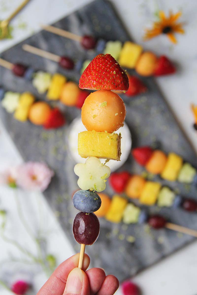Close-up of a rainbow fruit skewer held in a hand, showing the different types of fruit and lime zest garnish.
