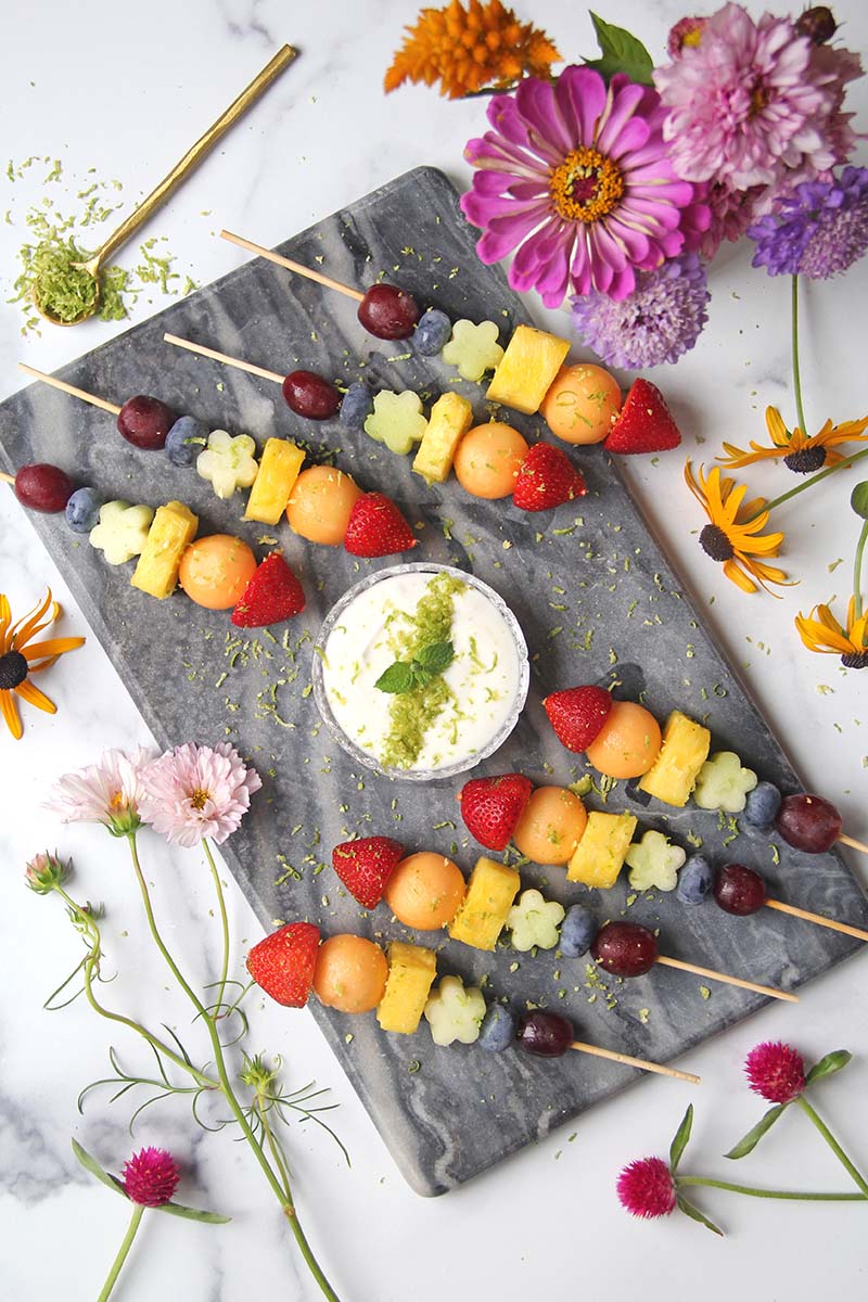 Overhead view of a platter of rainbow fruit skewers, shown alongside a vase of summer flowers.