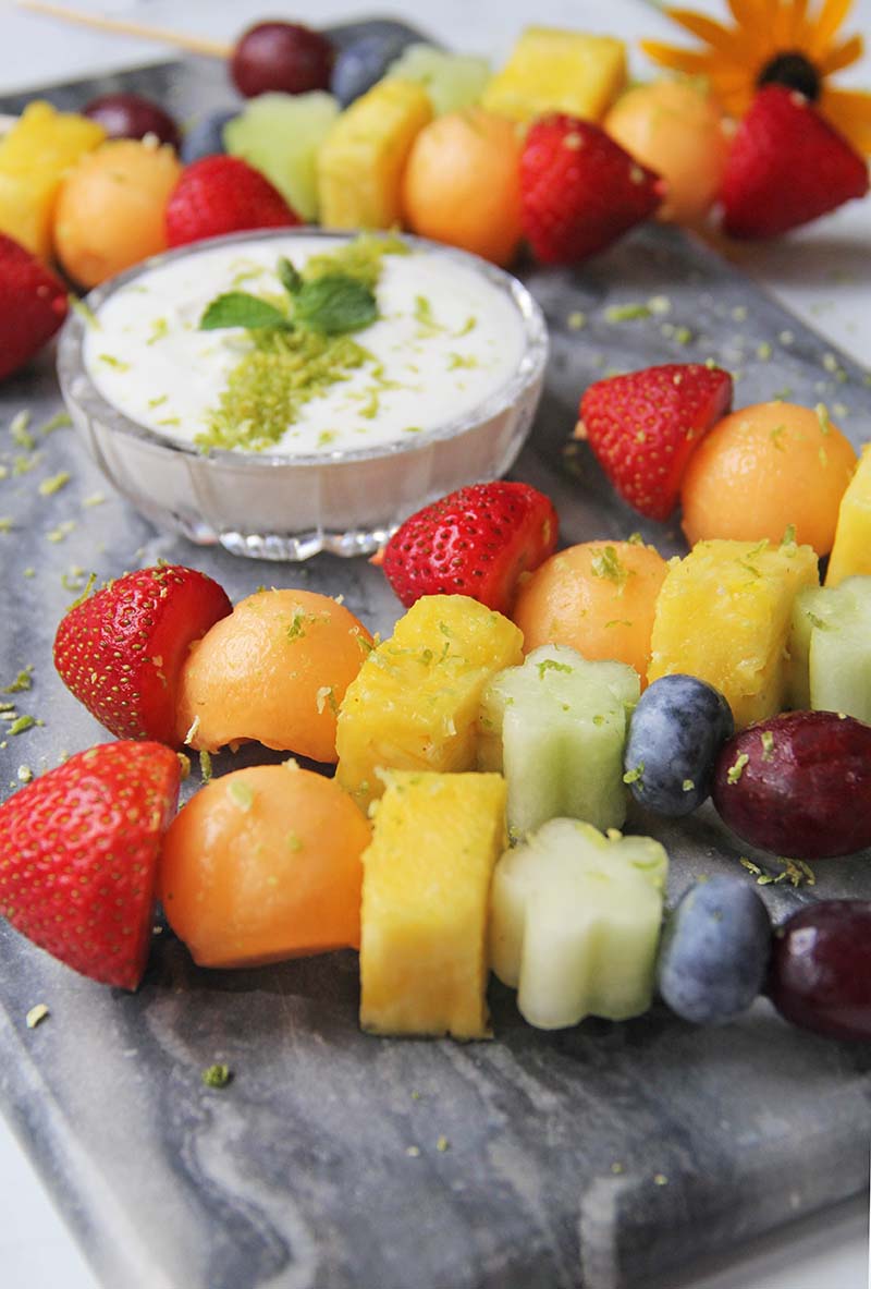 Angled view of rainbow fruit skewers and a bowl of mojito yogurt sauce, all garnished with a sprinkle of lime zest.