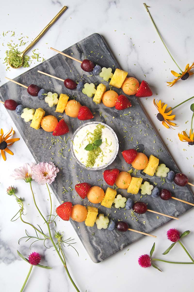Serving platter of rainbow fruit skewers on a white marble countertop, shown with summer flowers and a spoonful of lime zest.