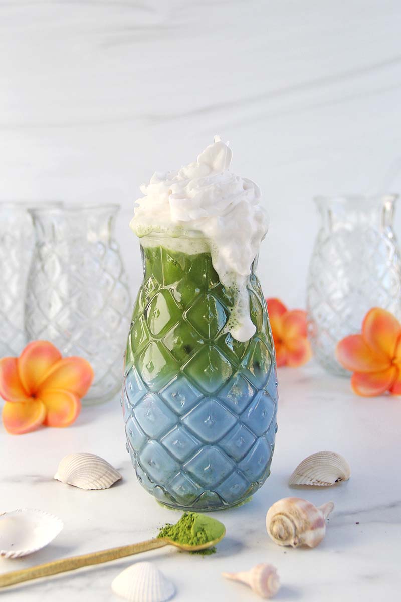 Blue matcha latte made with blue spirulina, topped with whipped cream, which is running down the side of the glass.