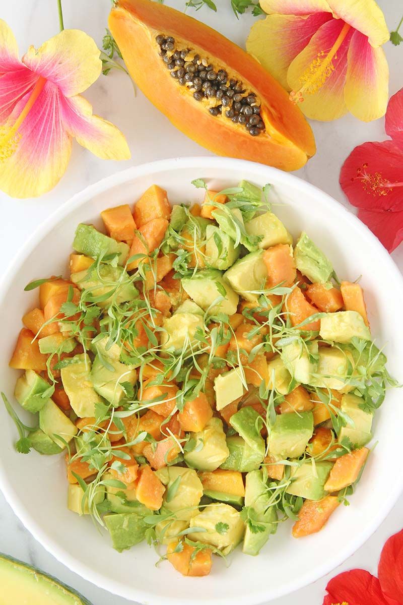 Bowl of tropical avocado papaya salsa shown from above on a marble countertop next to a papaya half and tropical flowers.