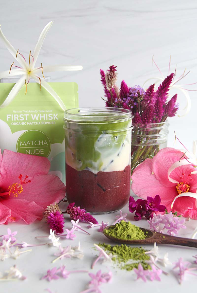 Iced Açaí Matcha Latte in a jar, shown with a bag of Matcha Nude matcha and tropical flowers.
