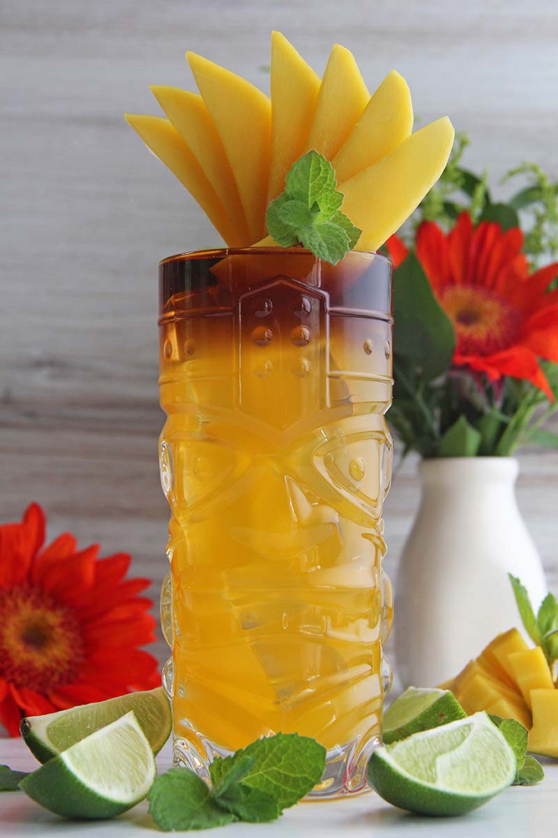 Mango rum punch in a tiki glass, garnished with a fan of mango slices and fresh mint, shown with Hawaiian flowers.