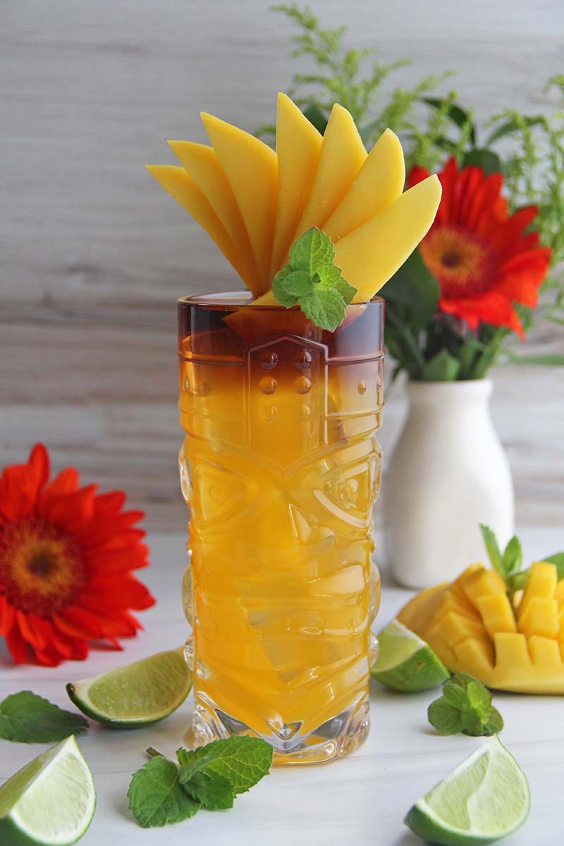 Tropical mango rum punch cocktail, shown next to colorful flowers, lime wedges, and a mango half.