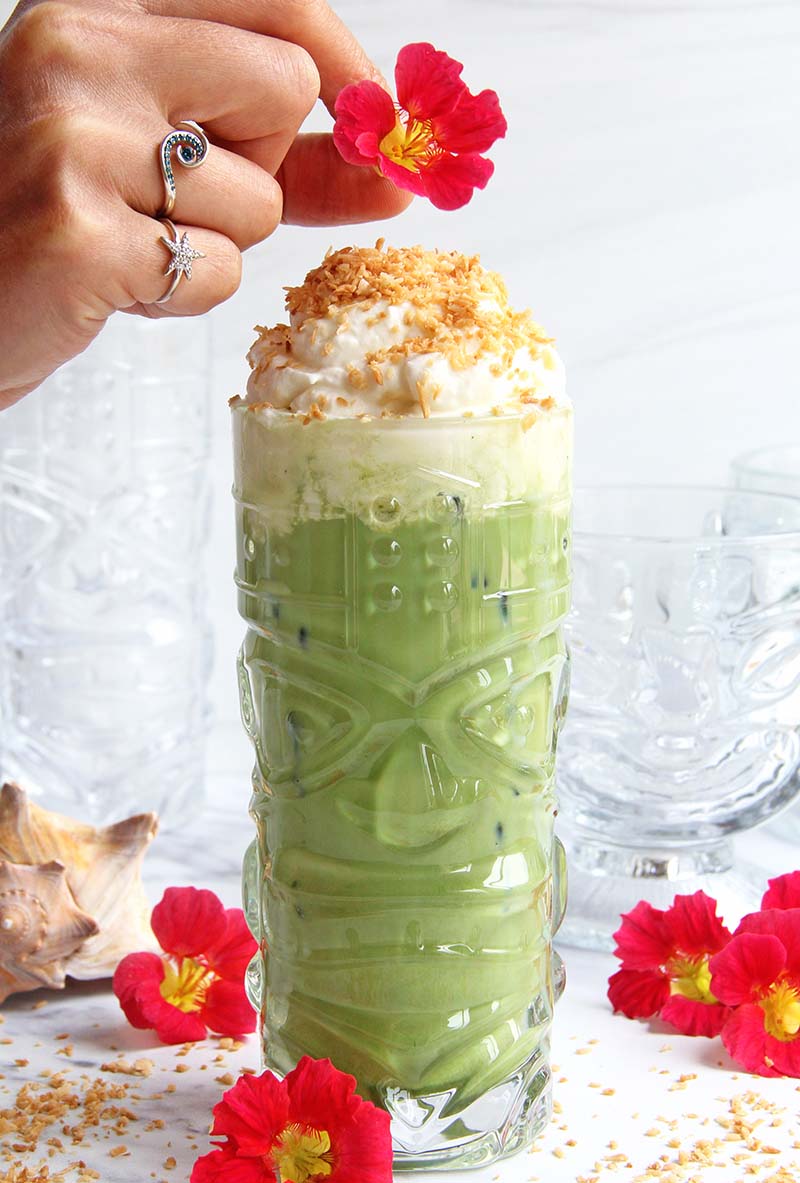 Woman's hand adding an edible tropical flower on top of a Toasted Coconut Matcha Latte in a tiki mug.