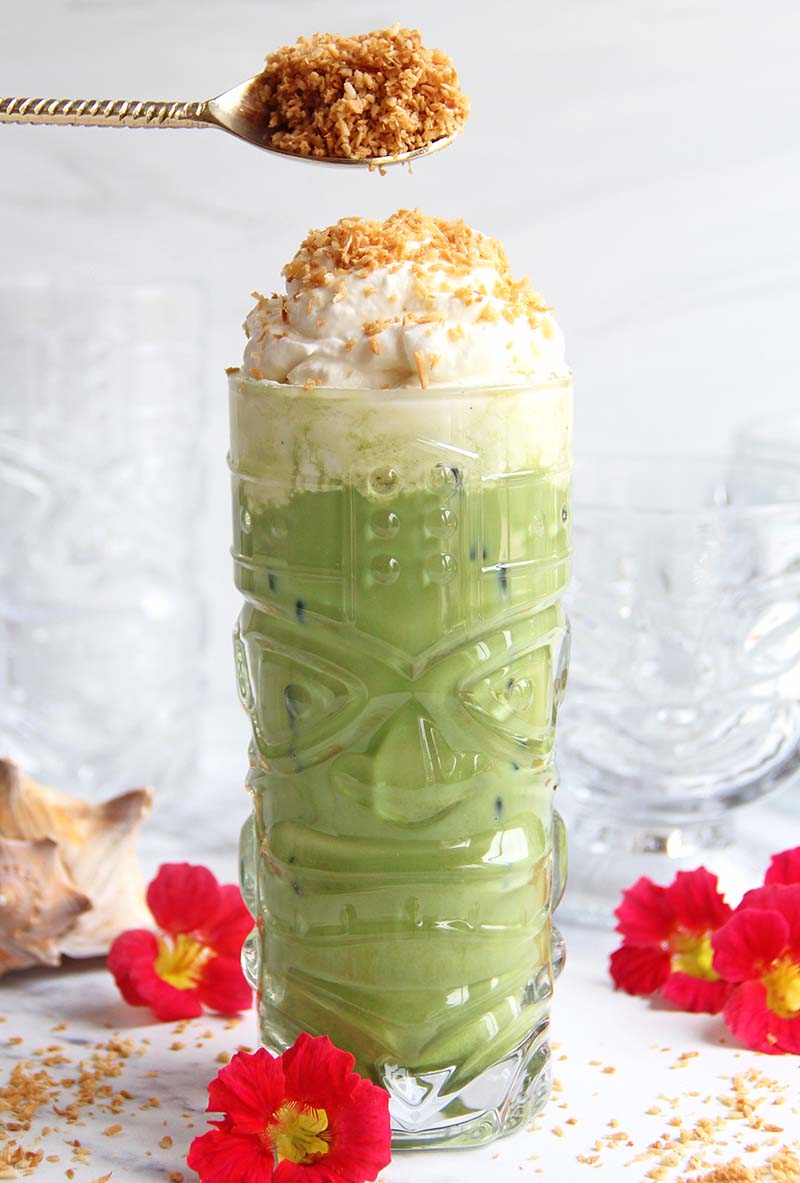 Spoonful of toasted coconut being sprinkled on top of a Toasted Coconut Matcha Latte in a white kitchen.