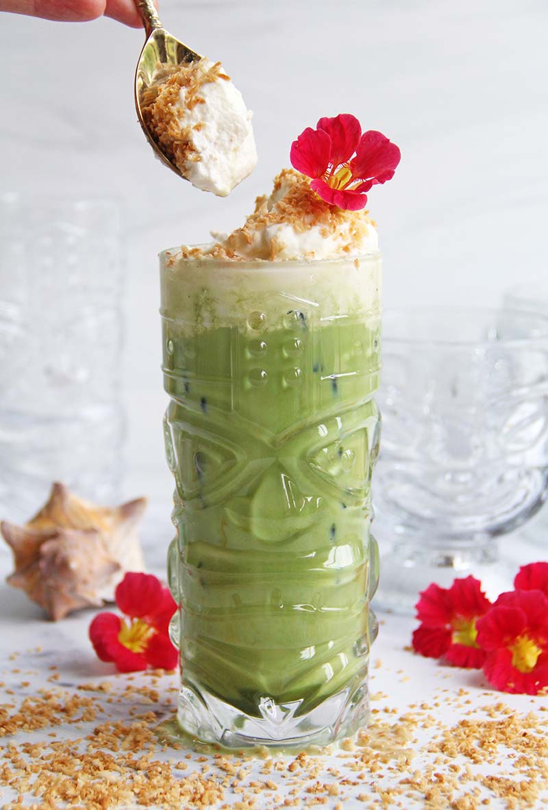Close-up of a spoon dipping into the coconut whip on a Toasted Coconut Matcha Latte, shown in a white kitchen with tropical seashells and flowers.