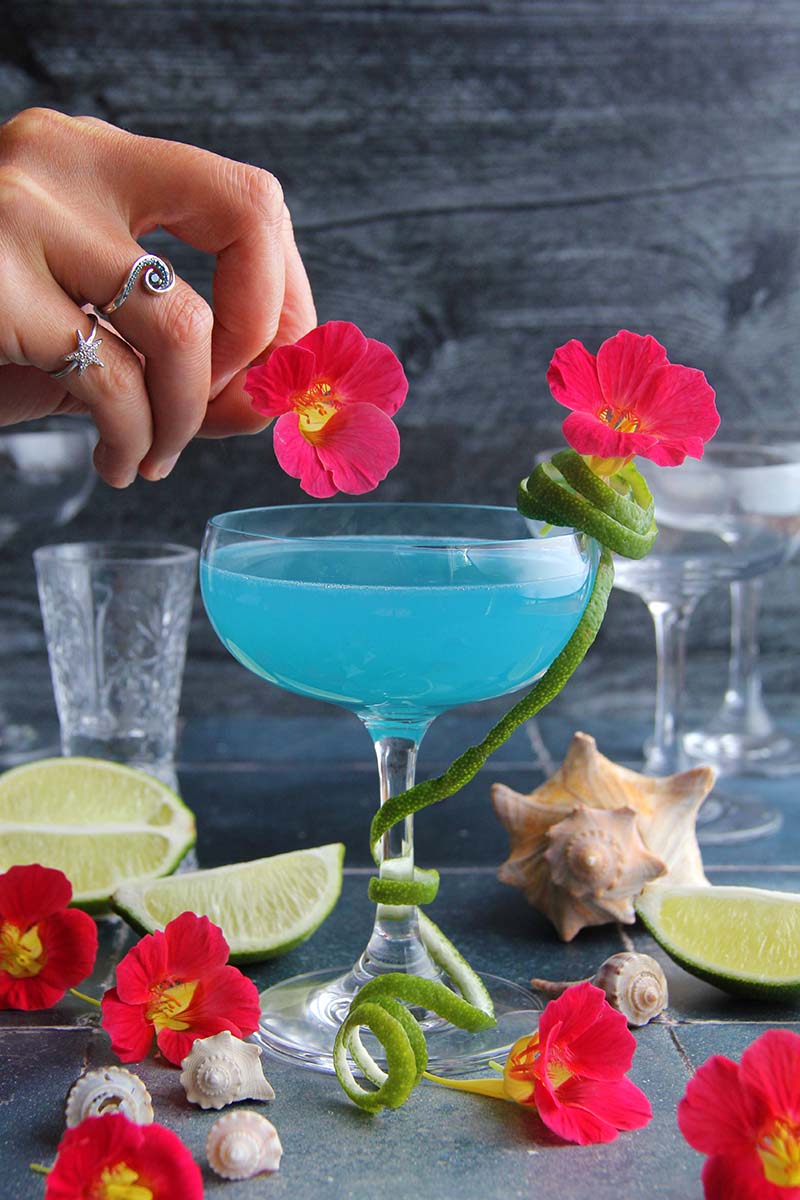 Woman's hand adding an edible tropical flower to a Blue Coconut Daiquiri cocktail.