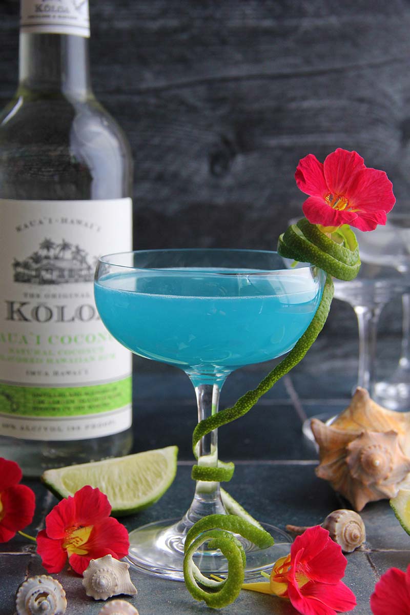 Blue Coconut Daiquiri, garnished with lime peel and an edible flower, shown next to a bottle of Koloa Rum coconut rum.