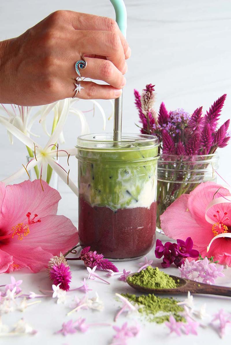 Woman's hand dipping a straw into a tropical acai matcha latte with purple (acai), white (soy milk), and green (matcha) layers.