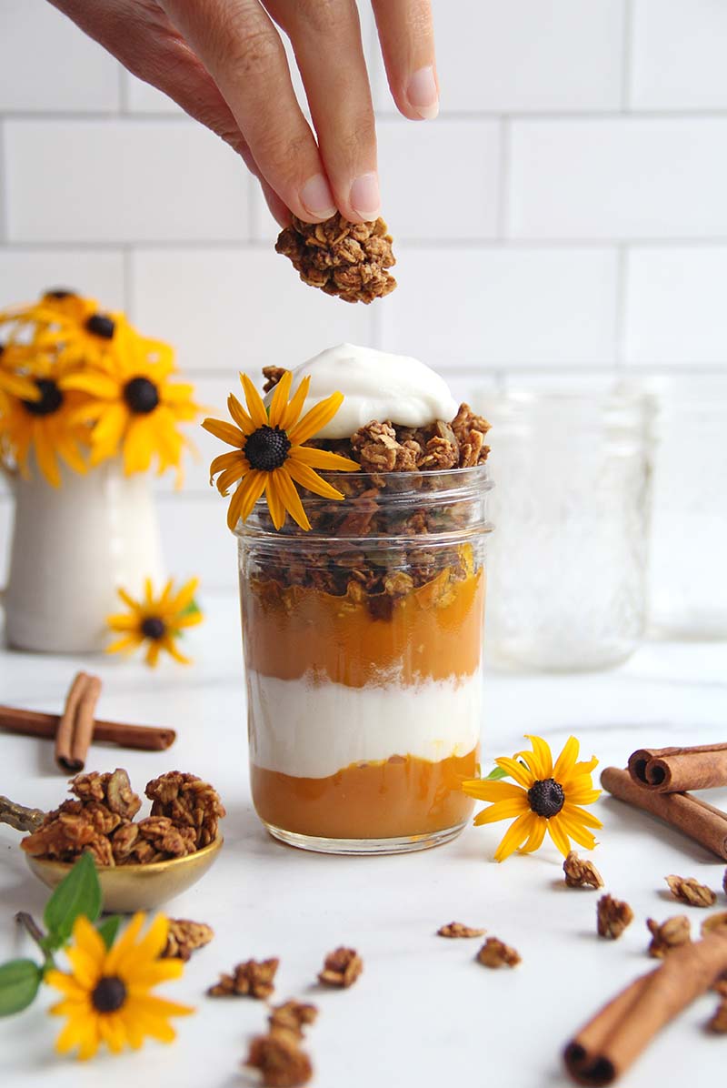 Woman's hand adding a chunk of granola on top of a pumpkin yogurt parfait in a mason jar.