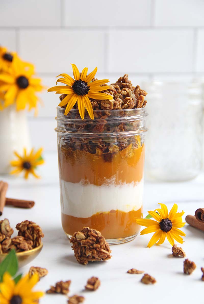 Close-up of a pumpkin yogurt parfait in a mason jar, shown in a white kitchen with fall flowers.
